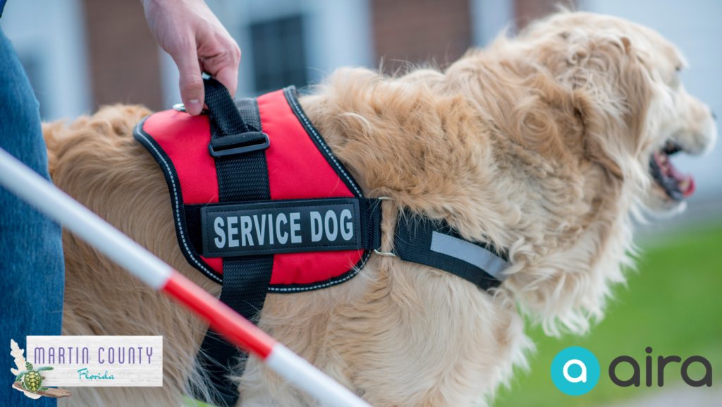 A hand holding the red harness of a guide dog, featuring the Martin County logo and Aira App logo